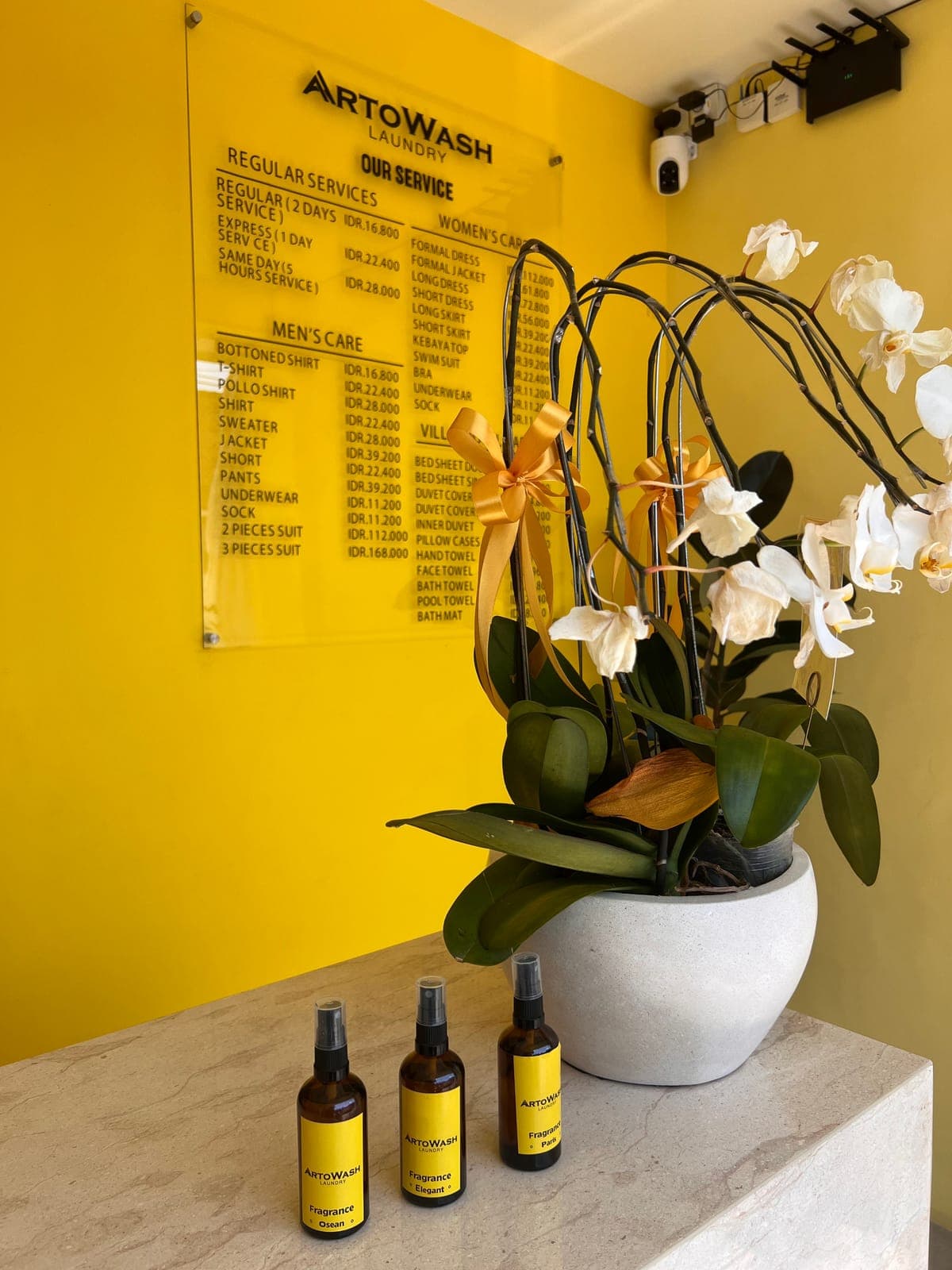 The service board on the yellow wall behind the counter, with ArtoWash fragrance sprays and an orchid
